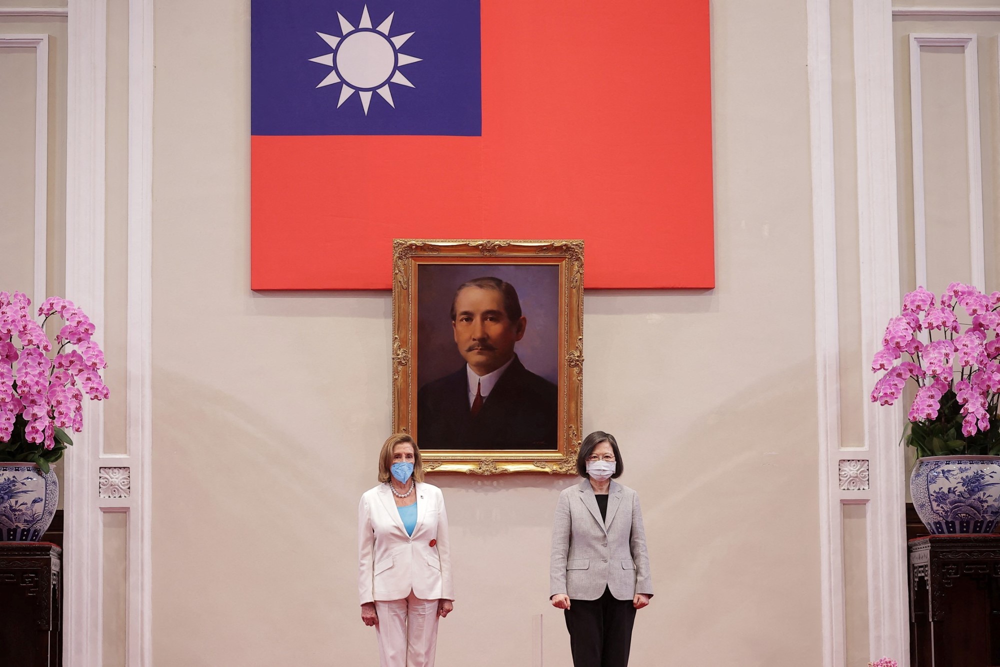 Two women wearing suits stand in front of a portrait and flag.