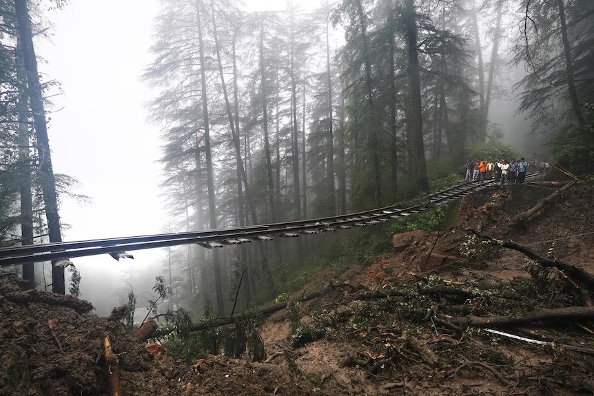 Part of a train track in a forest, which has separated from the ground beneath it due to a landslide. People stand on the side of it