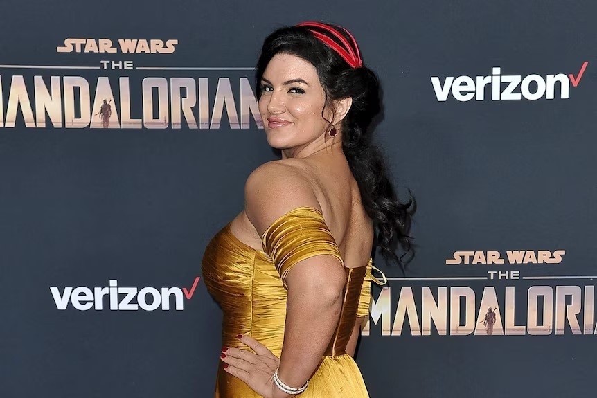 A Caucasian woman, with black hair, wearing a gold dress, posing in front of a sign with 'The Mandalorian' logo.