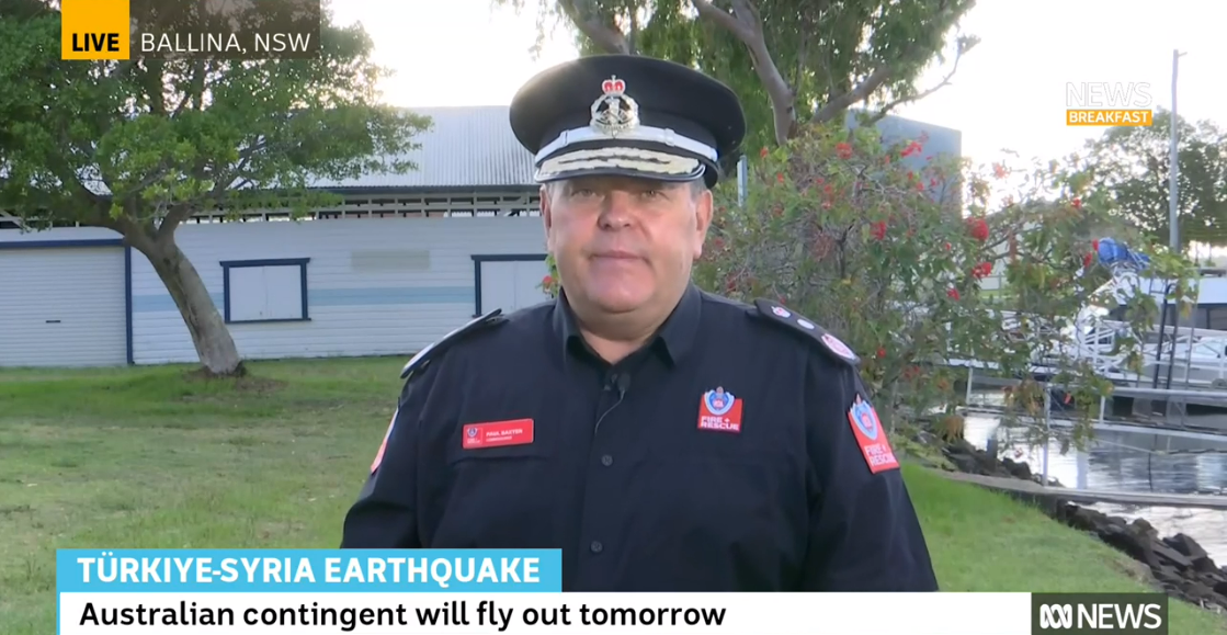 A man in an official's uniform and hat stands on a grass park being interviewed