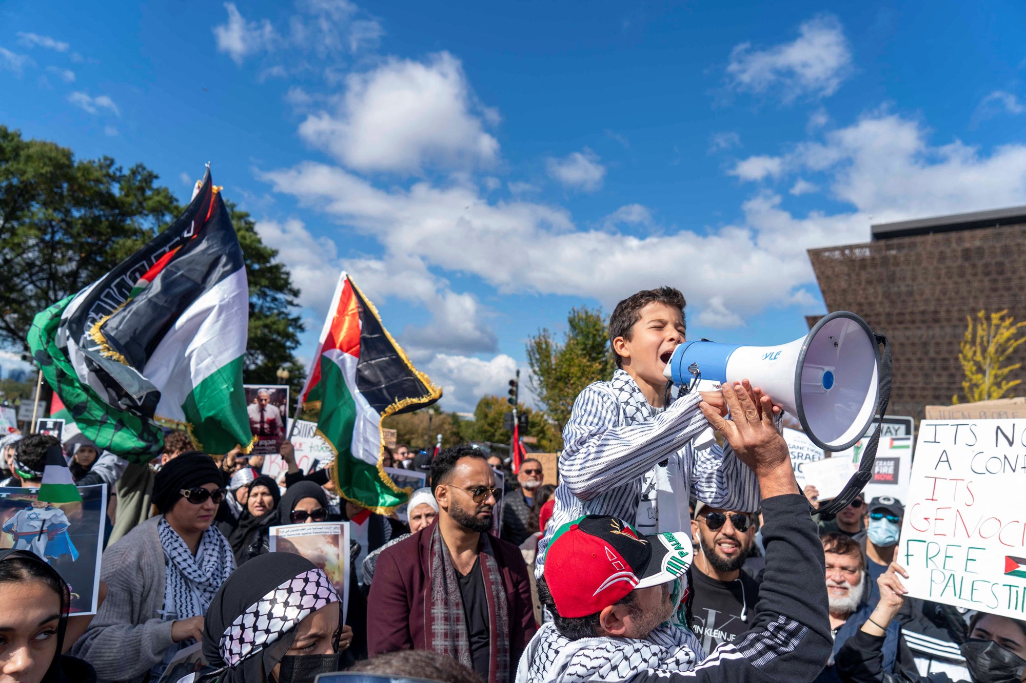 A boy speaks through a loudspeaker and People raise flags and posters during a rally held by American Muslims for Palestine calling for a cease fire in Gaza near the Washington Monument in in Washington