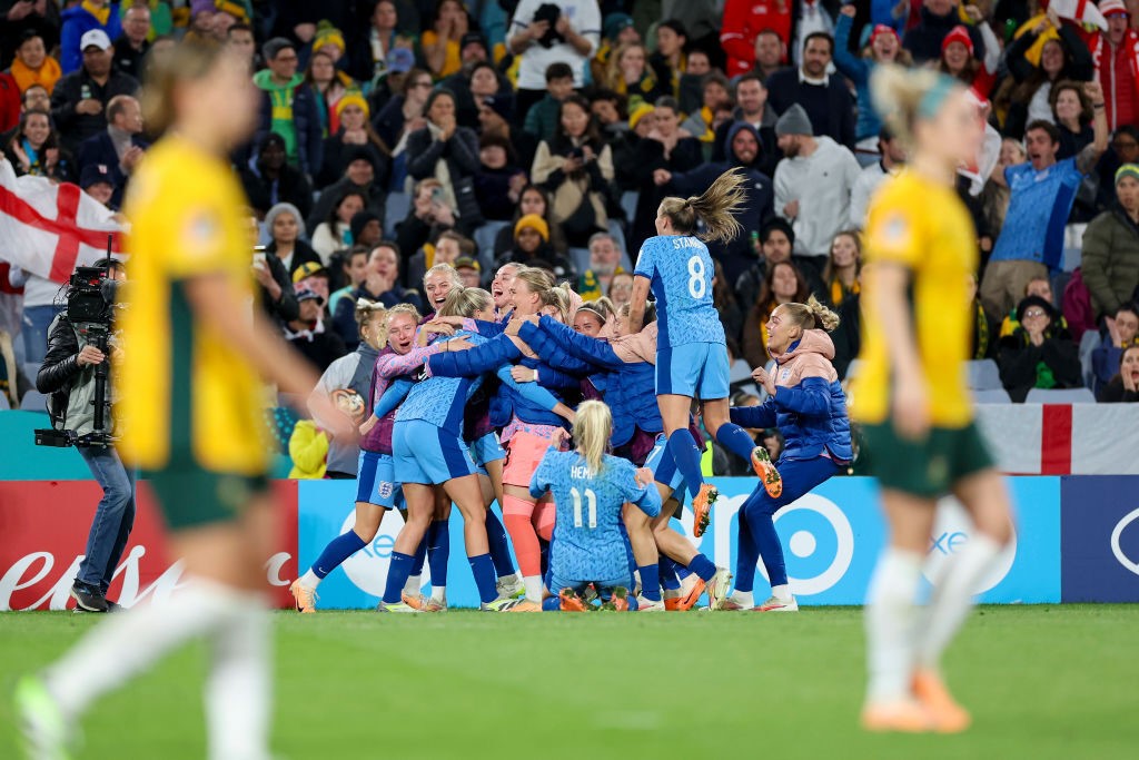 England players pile on in celebration as Matildas walk in the foreground.