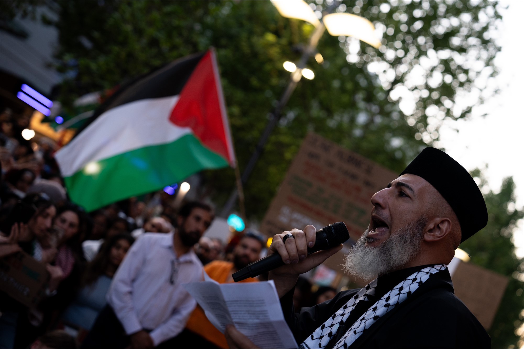 A man holding a microphone, a palestinian flag in the background. 