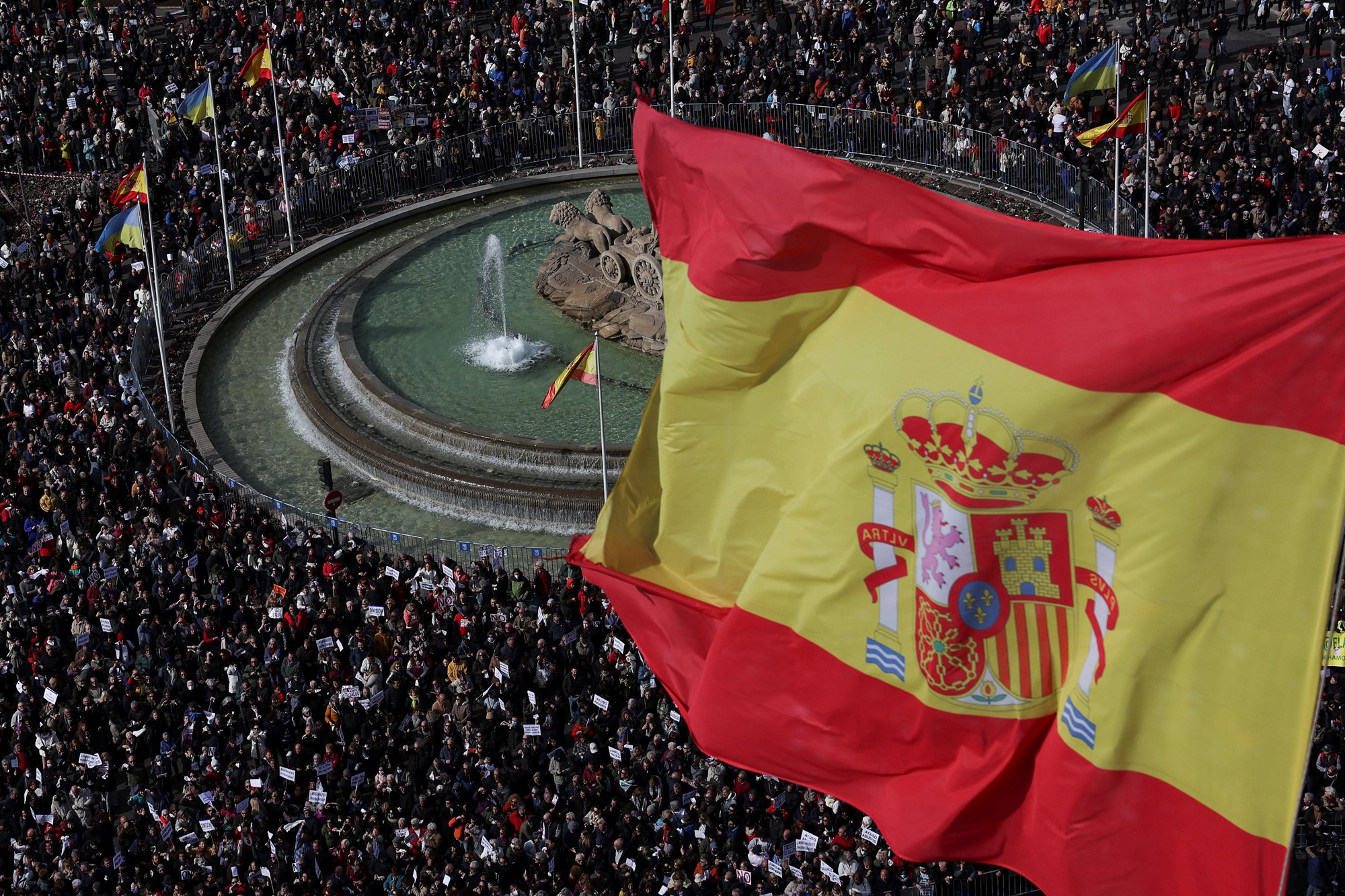 A crowd of people around a fountain beneath the flying Spanish flag