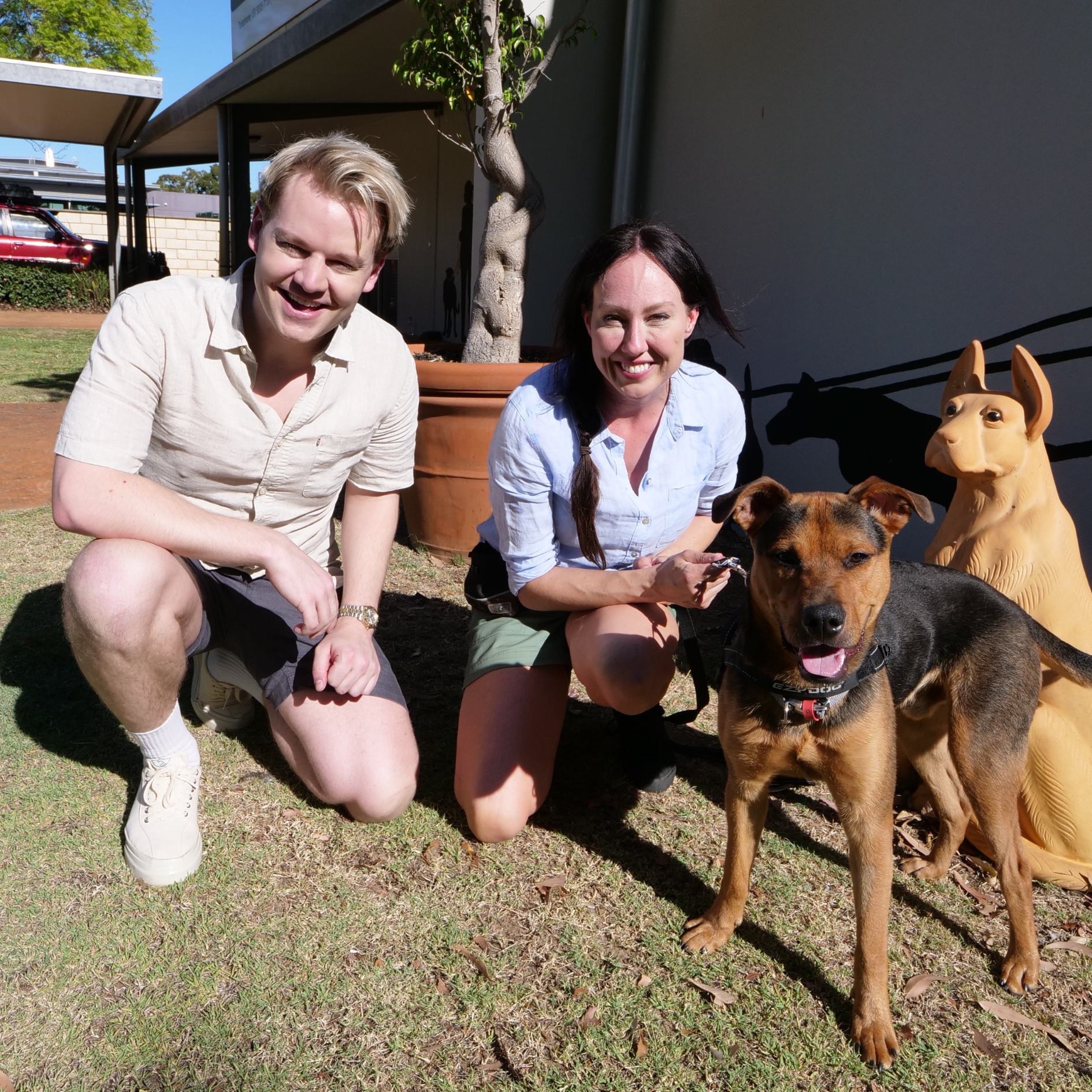 Joel Creasey and Laura Vissaritis kneel down beside a brown and black dog in the sun