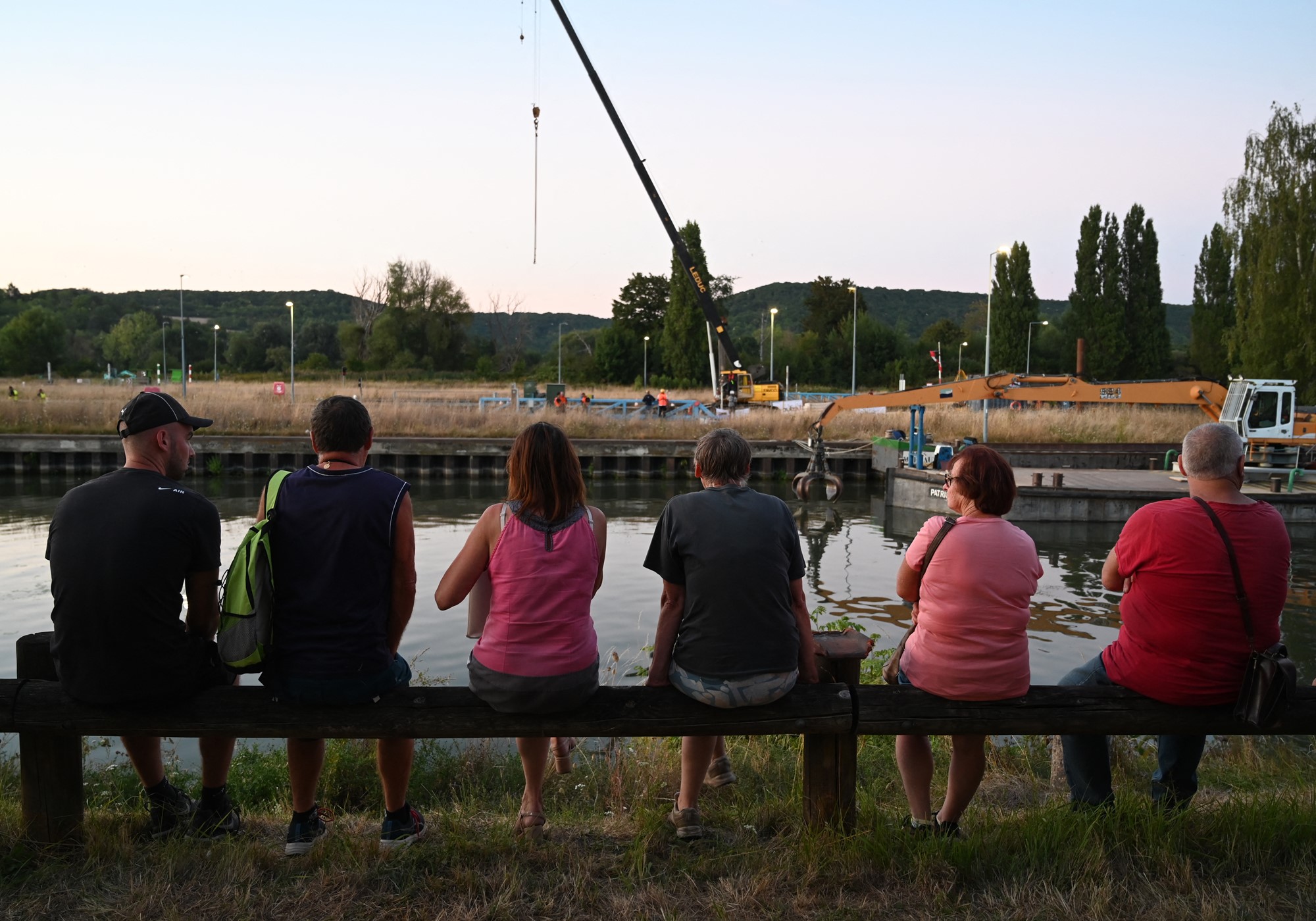 Onlookers sit and watch a crane over a river.