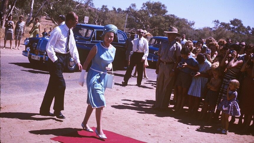 Queen Elizabeth dressed in a blue dress and hat and gloves walks the red carpet with her husband in front of a crowd of people.
