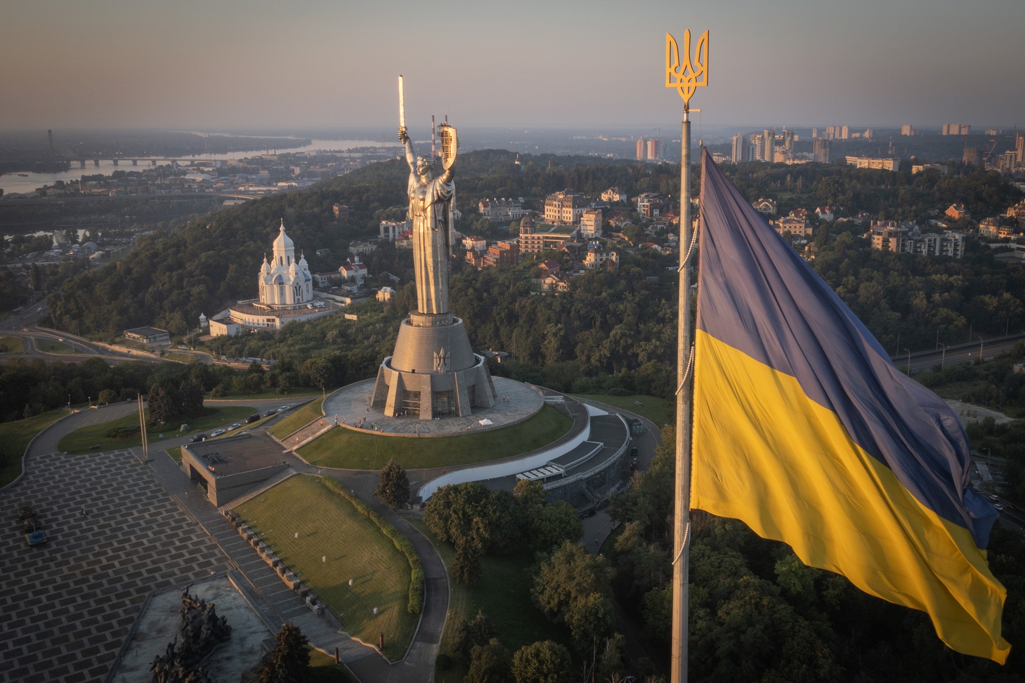 A wide photo of a tall statue of a woman holding a sword and shield. In the foreground is a tall Ukrainian flag