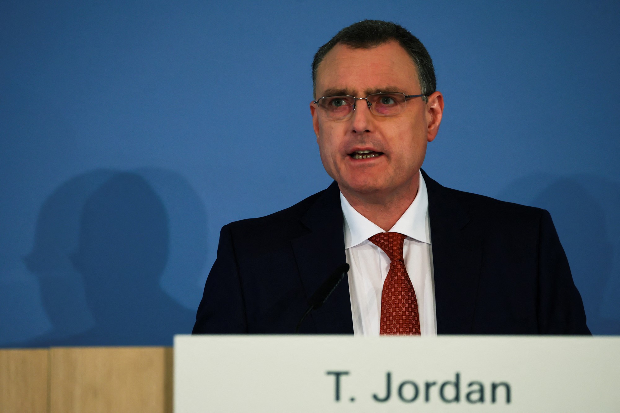 A middle aged man wearing a suit jacket with a white shirt, red tie and glasses speaks while standing behind a lectern and in front of a blue wall.