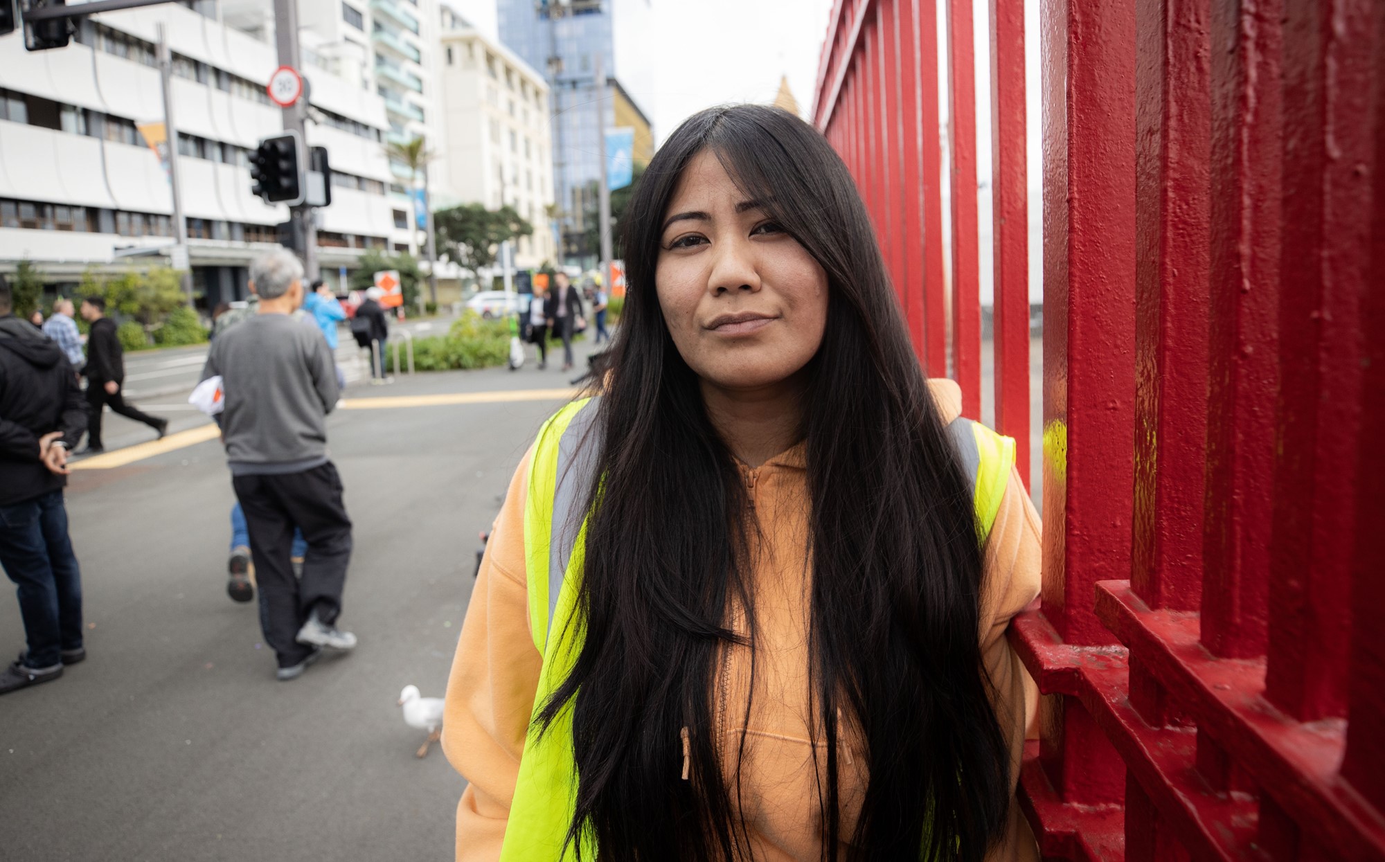 a woman with long black hair, orange jumper and high vis vest stands in front of busy street 