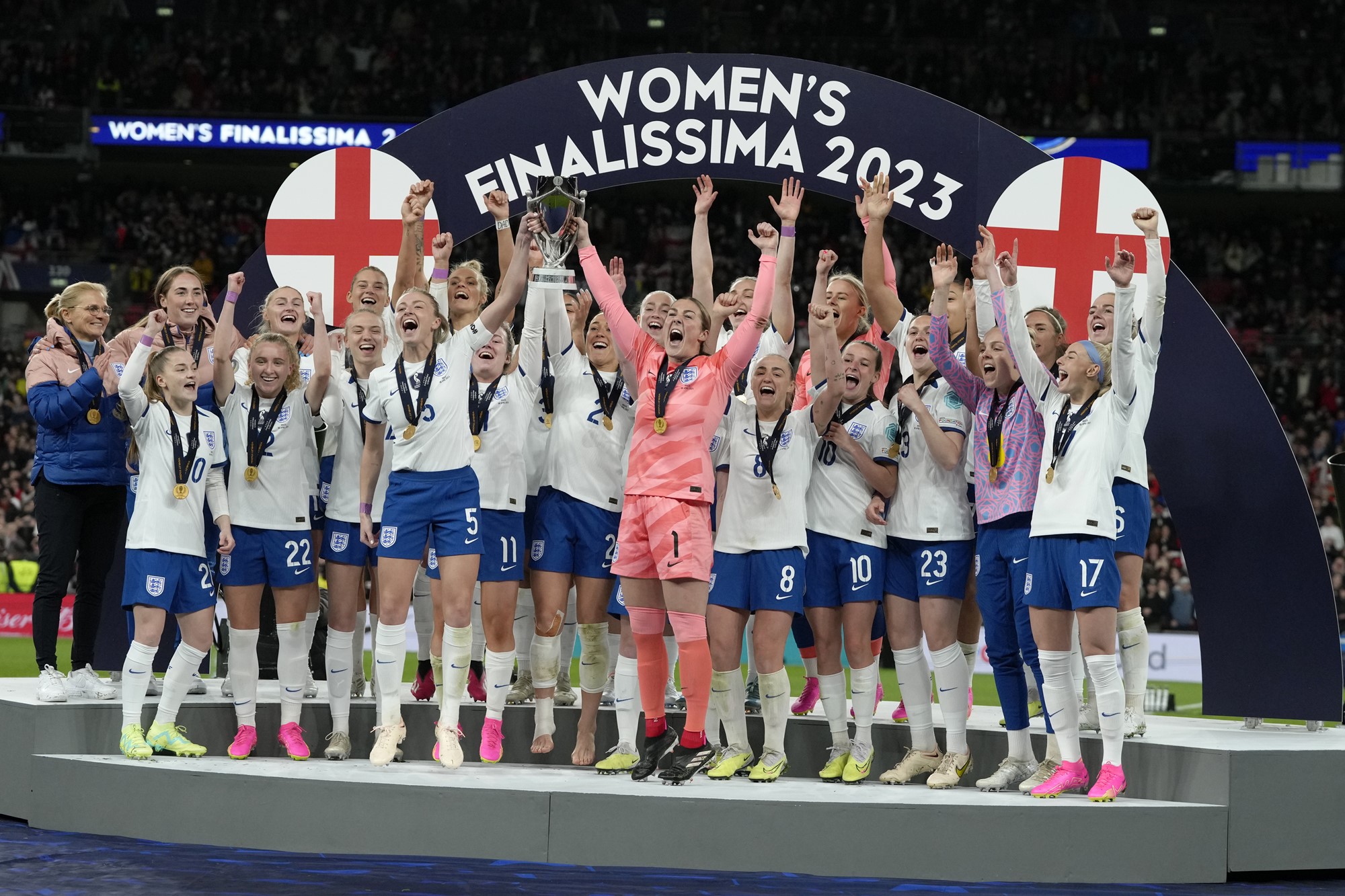 The England women's team chears holding a trophy under teh Women's Finalissima 2023 banner. 