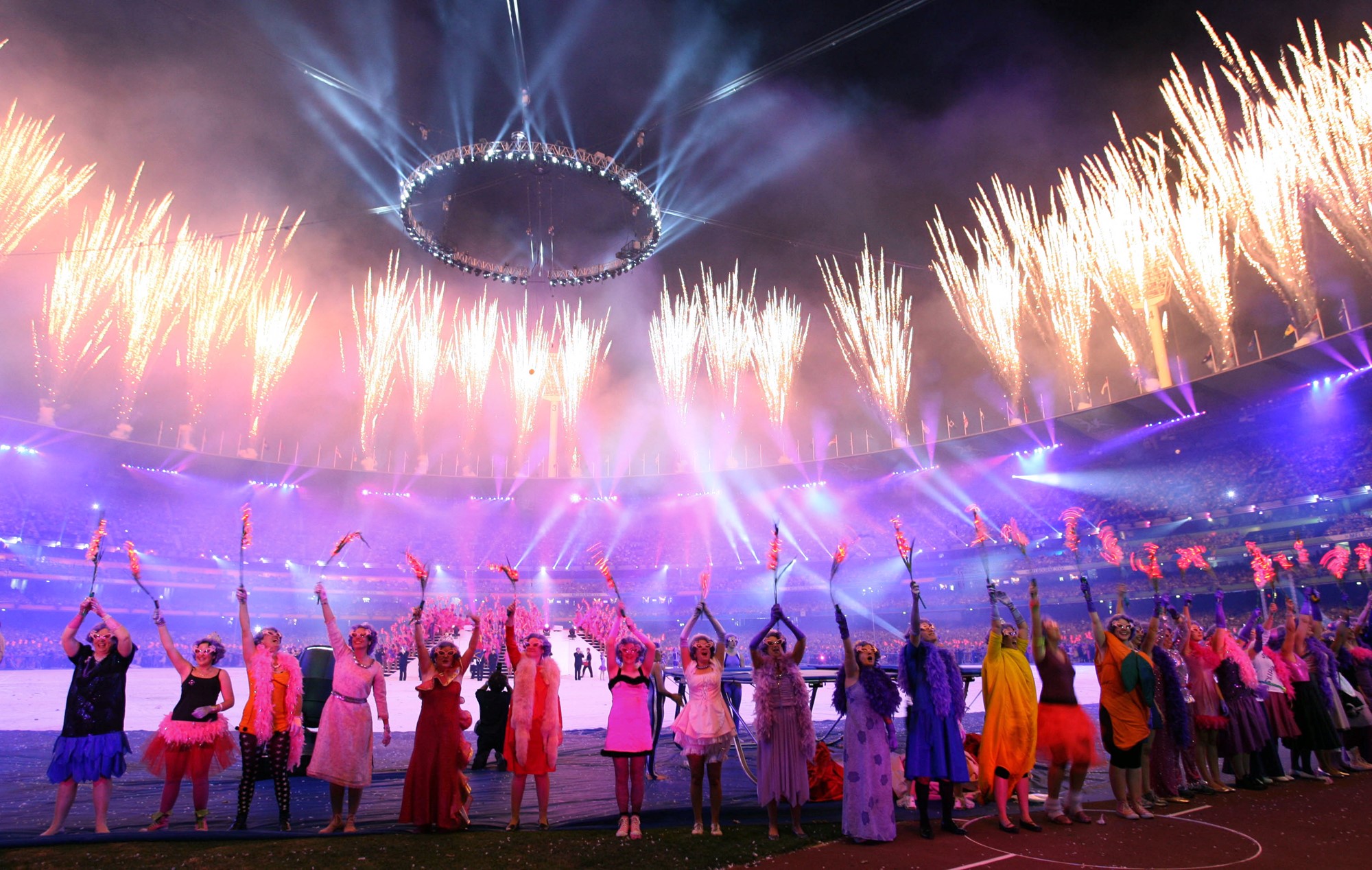 Dancers in purple wigs and Dame Edna glasses cheer as fireworks go off around the roof of a stadium. 