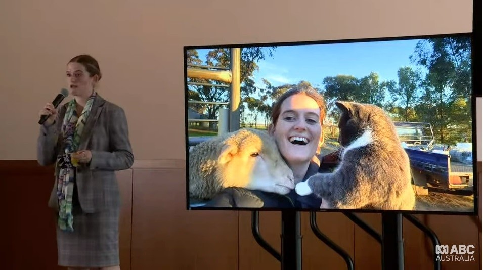 A woman in a plaid suit holds a microphone. A screen next to her shows a picture of her with a lamb and cat.