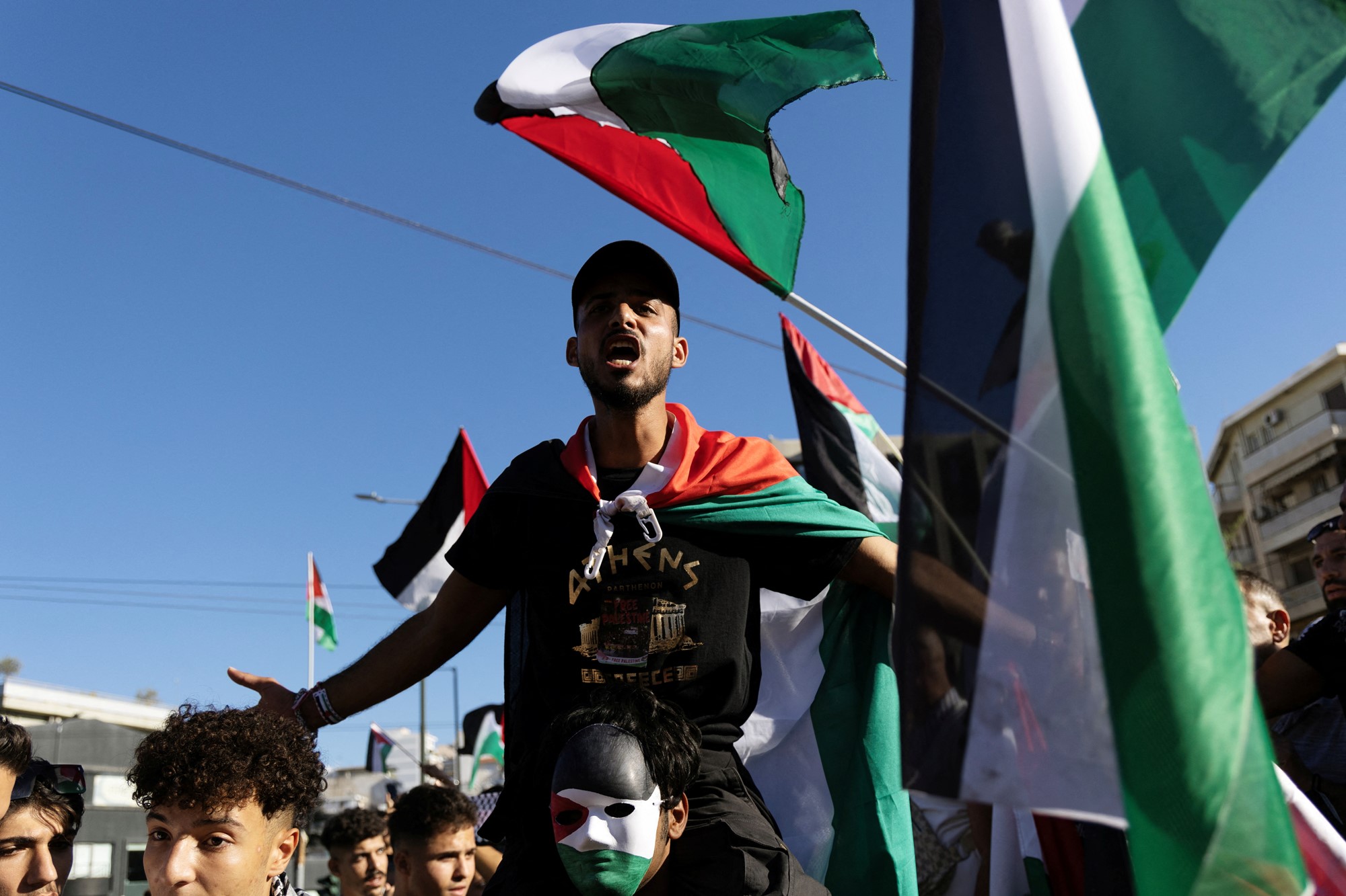 A man sits on another's soldiers with his arms spread wide, shouting with his mouth open. Palestinian flags are waved around him