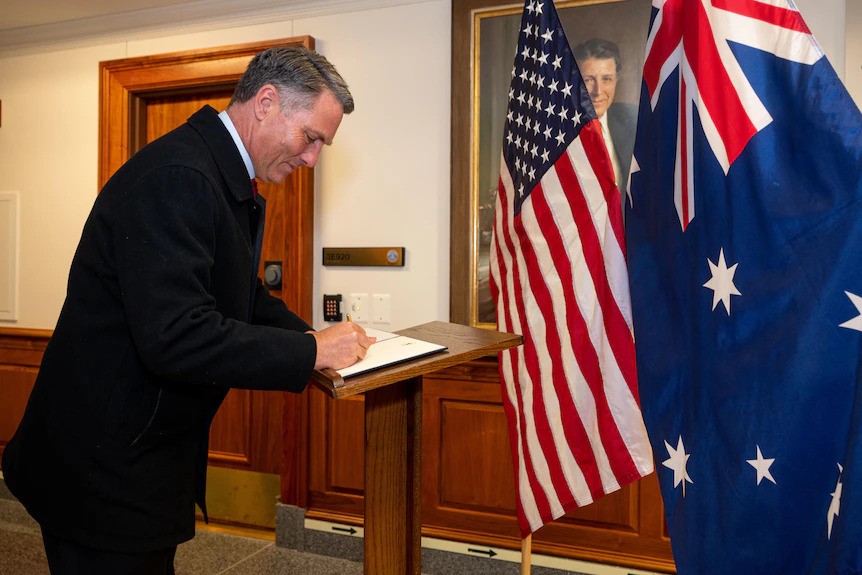 Richard Marles signing paper at a lecturn next to large Australian and US flags