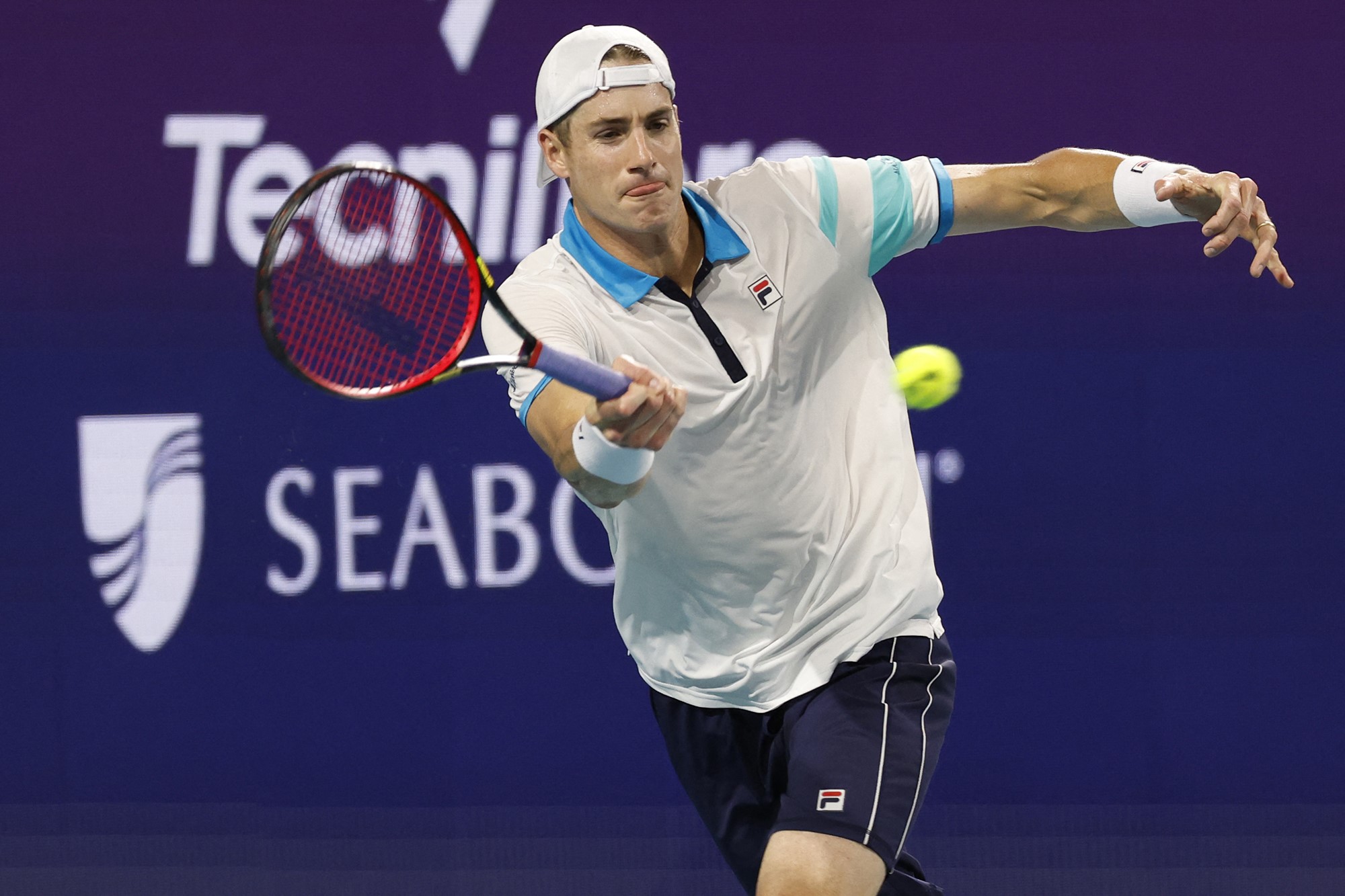 A man in a white tennis polo and backwards hat hits the ball 