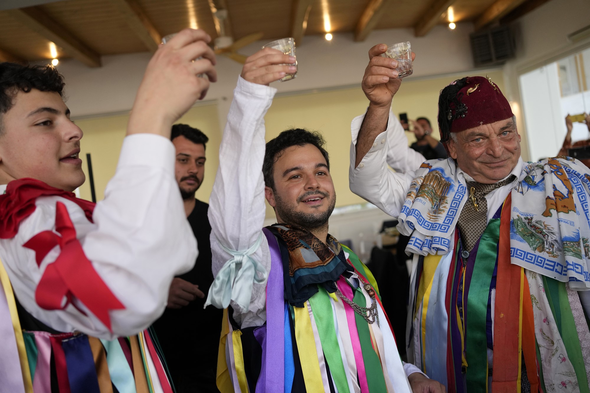 People in traditional ribboned Greek folk costumes cheer with small glasses.