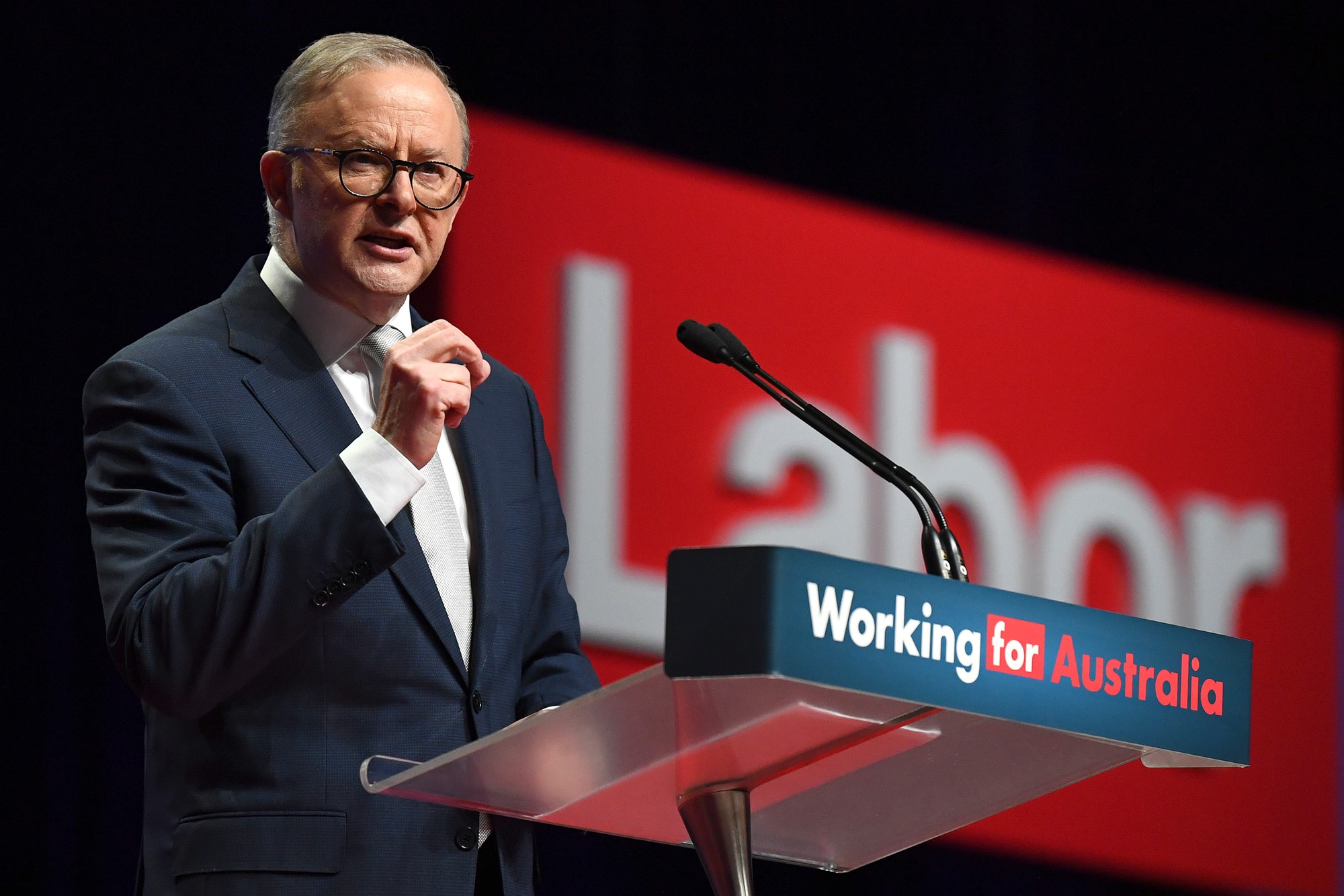 A man wearing glasses and a suit stands behind a lectern labelled "Working for Australia".