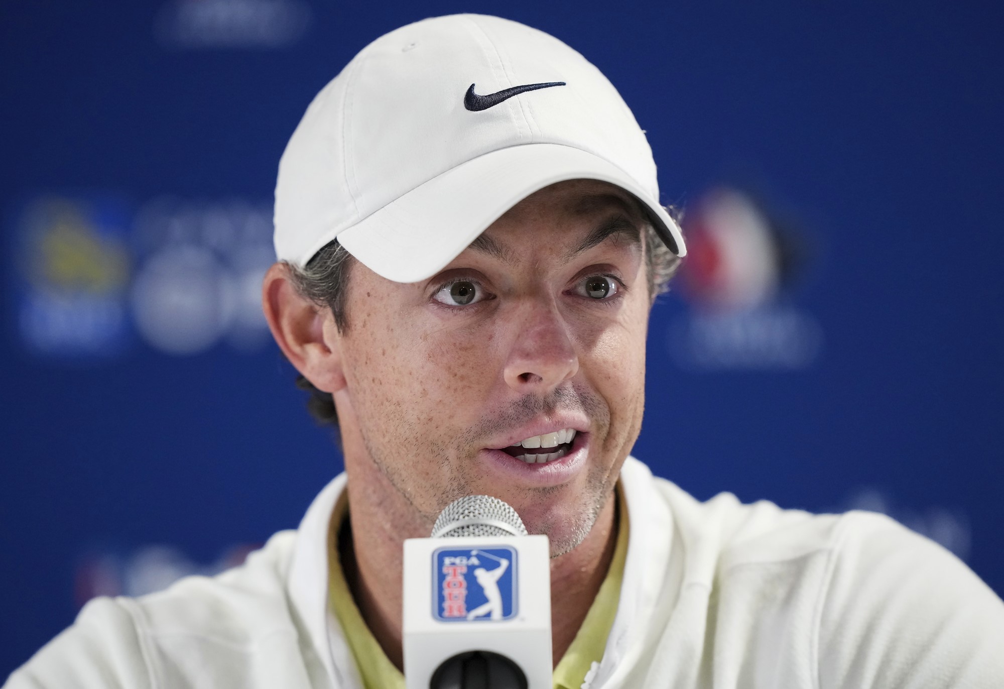 Rory McIlroy speaking into a microphone. He's wearing a white cap and white sweater. He's got short brown hair and brown eyes.
