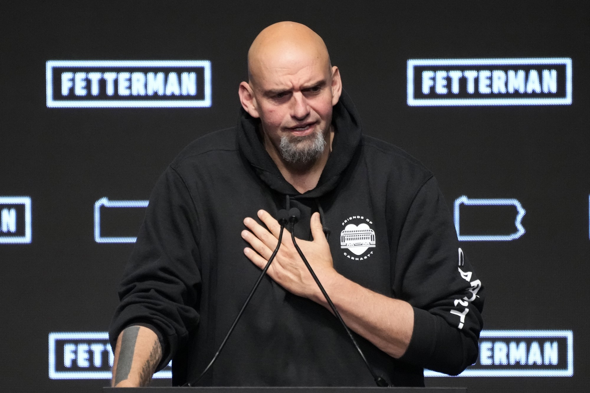 A tall, bald man with grey beard stands in front of black and white FETTERMAN signs, holding his hand to his chest