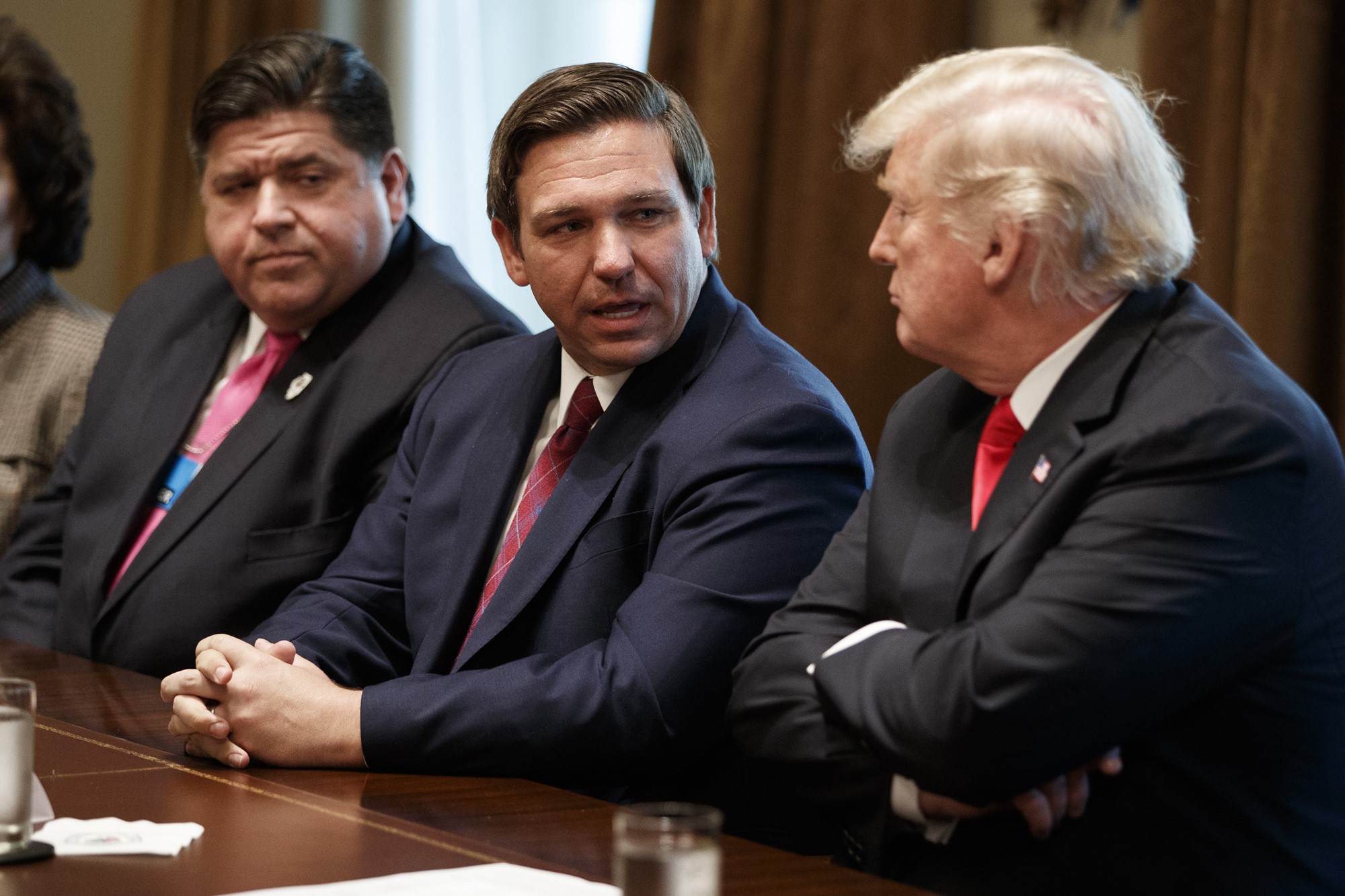 Three men, one with white hair, sit next to each other at a table, wearing suits and ties