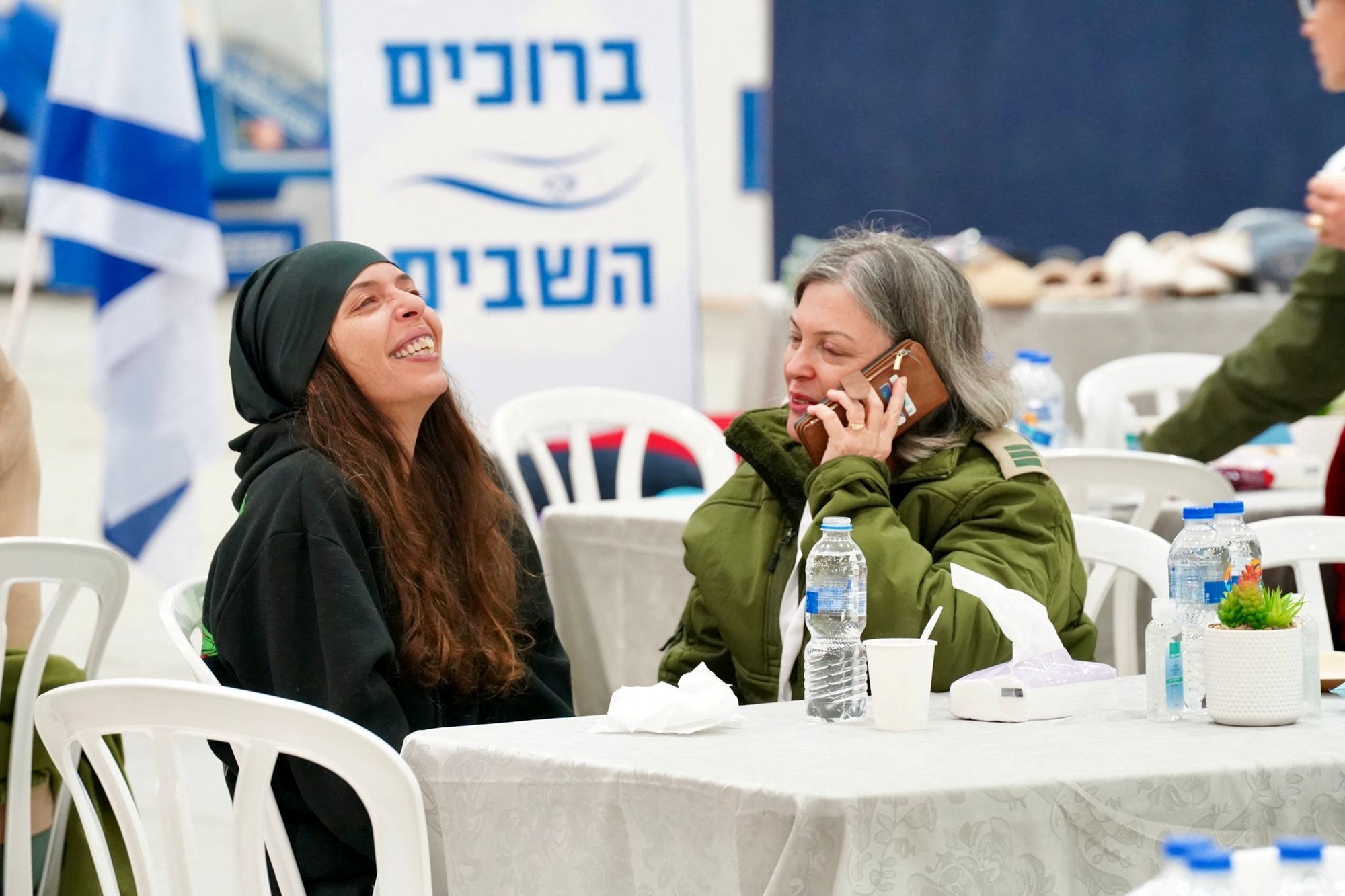 A woman looks up an laughs, another is speaking on the phone as they sit down at a table