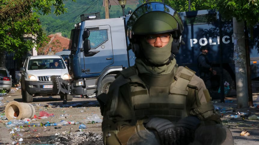 A soldier golds a gun and stands on a road covered with glass and litter. Some vehicles in the background, including a police truck