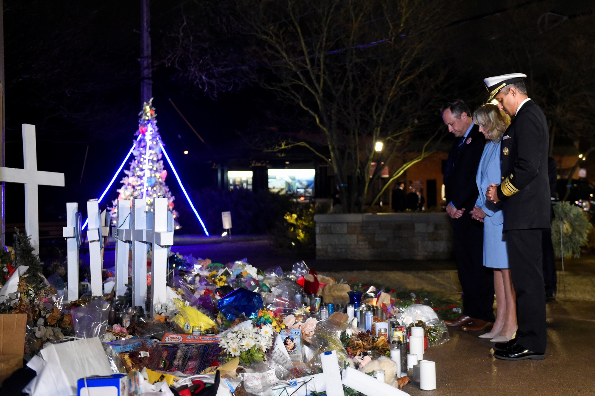 Three people stand in front of floral tributes.