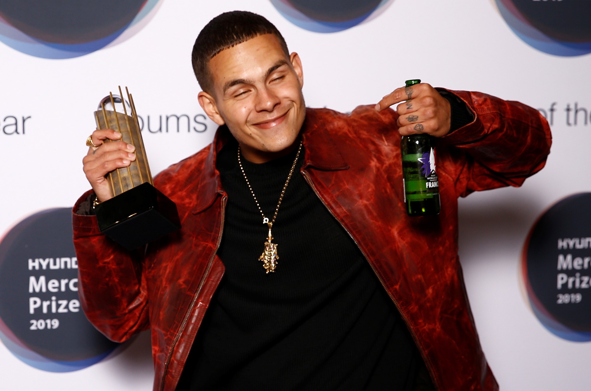 A man holding an award and a beer, smiles next to a Mercury Prize press backdrop