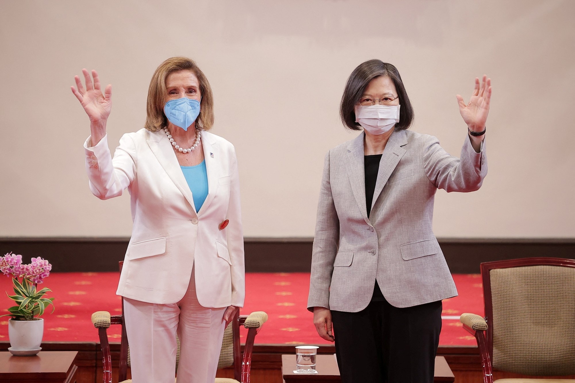 Two women wearing suits and face masks wave.