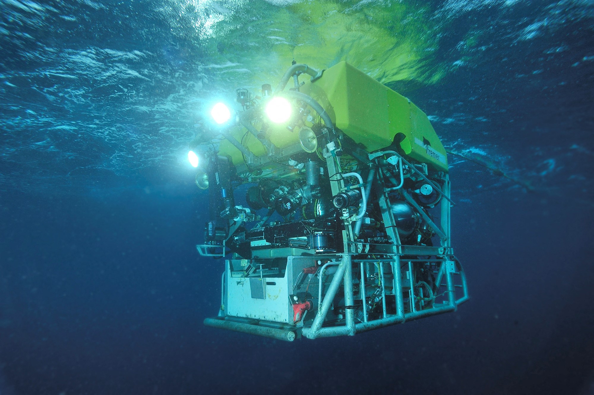 A cubic bright yellow and silver mass of beams and pipes, floating underwater with bright headlights.