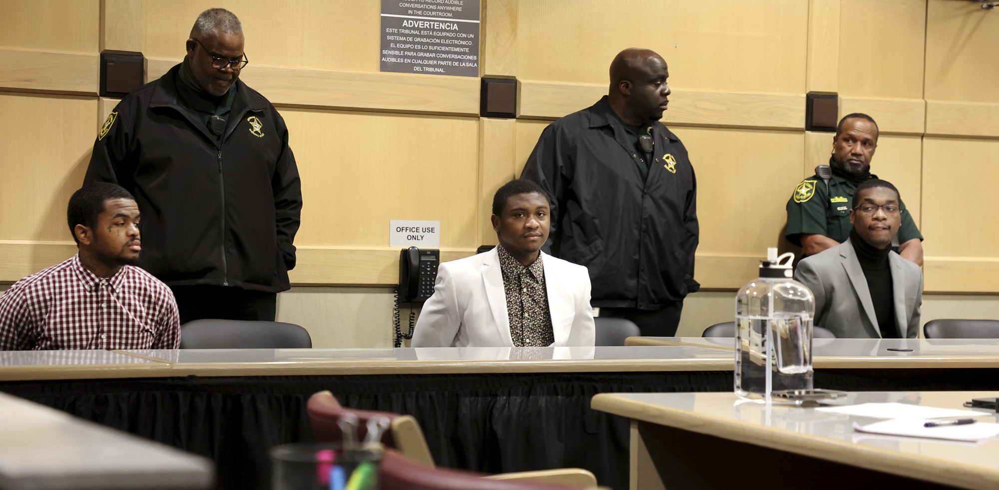 Three men sit at a court table with guards behind them. 