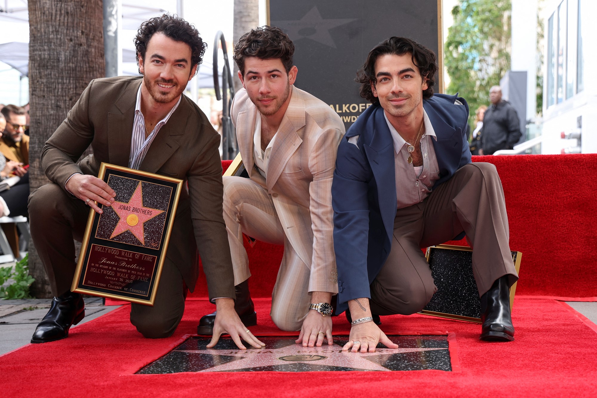 Three young men with dark hair and beards wearing suits kneel down pressing their hands on a star on the Hollywood Walk of Fame surrounded by red carpet.