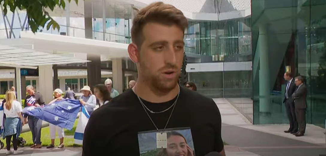 a man with a t shirt with a picture of a hostage on it talks to camera out front of parliament house