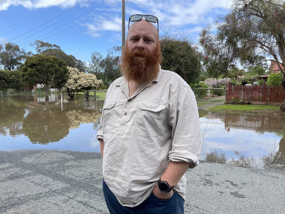 A bald man with a red beard stands in front of floodwaters.