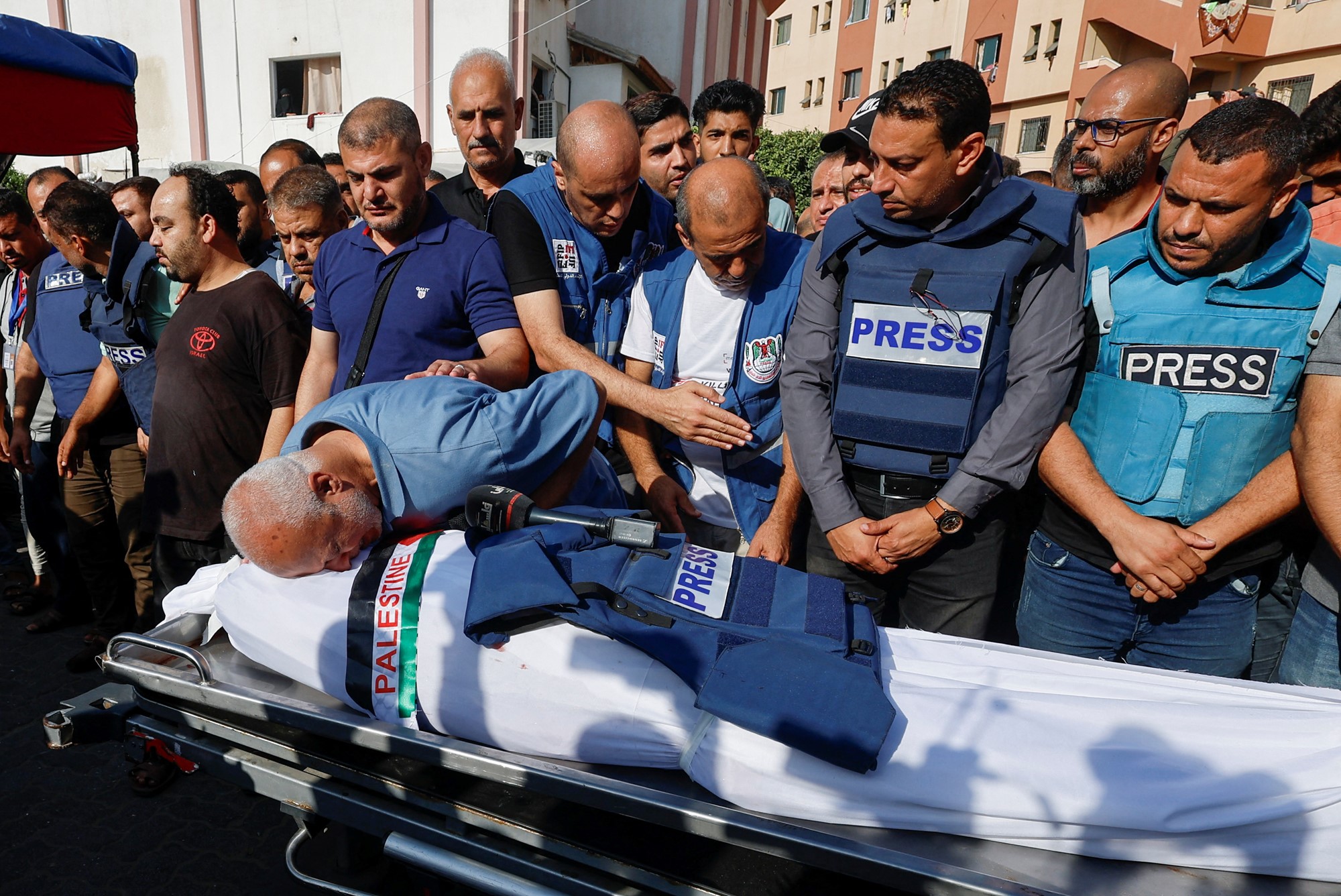 Journalists wearing press vests stand over white sheeted body of journalist wrpped in Palestinan badge. 