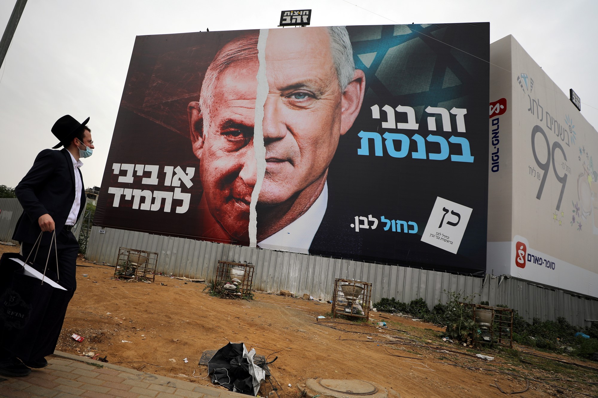 An Orthodox Jewish man stands beside a large billboard showing the faces of Benjamin Netanyahu and Benny Gantz, split down the middle.