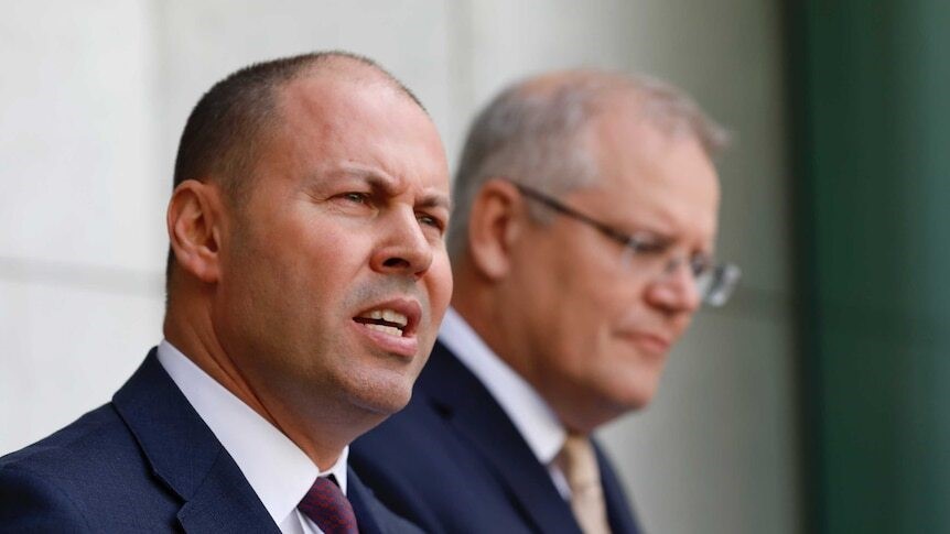 A close up of Frydenberg speaking at a press conference with Morrison behind him, out of focus.