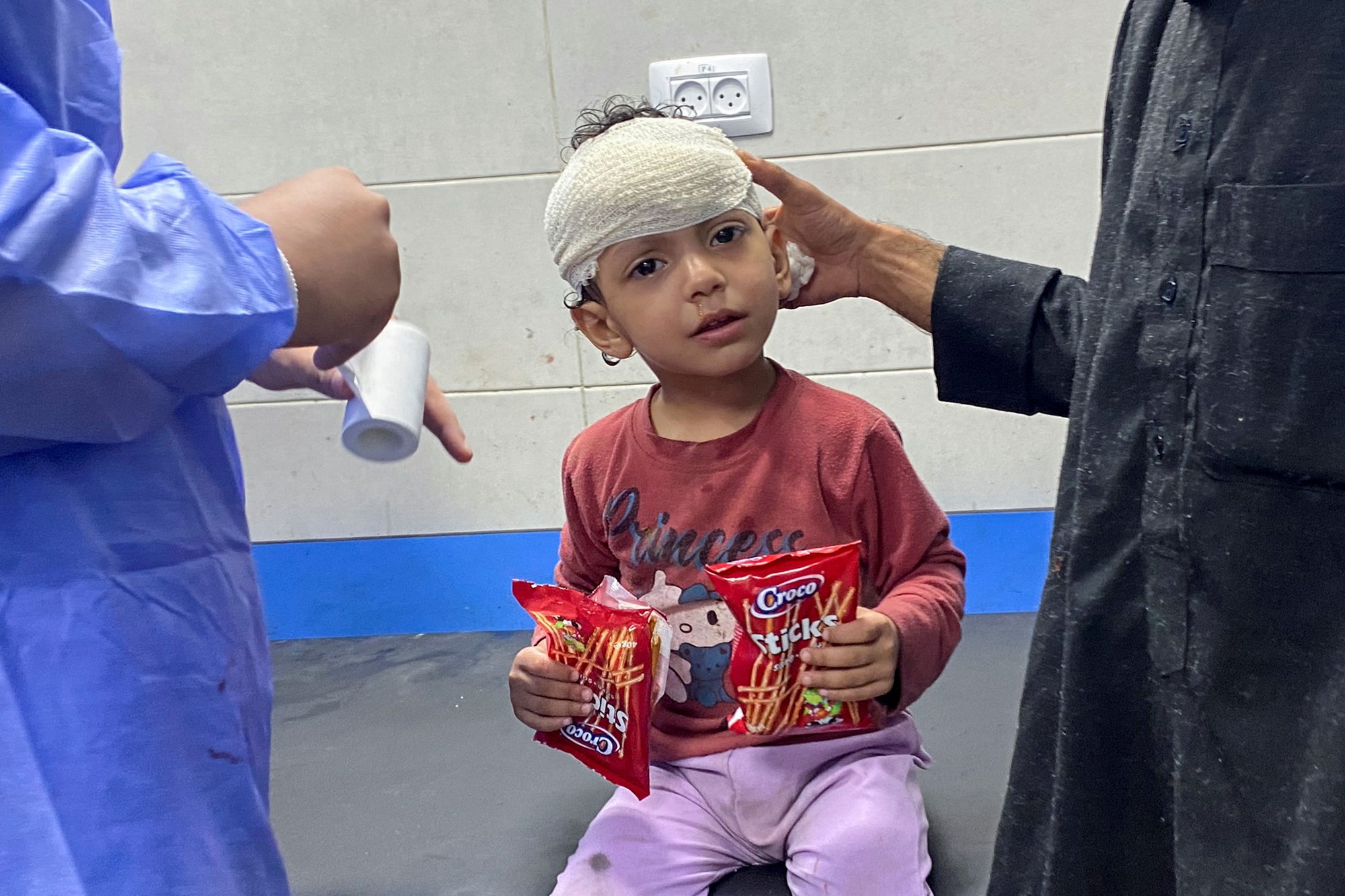 Image of a little girl, in her hands are two bags of pretzel sticks, she has a bandage around her head.