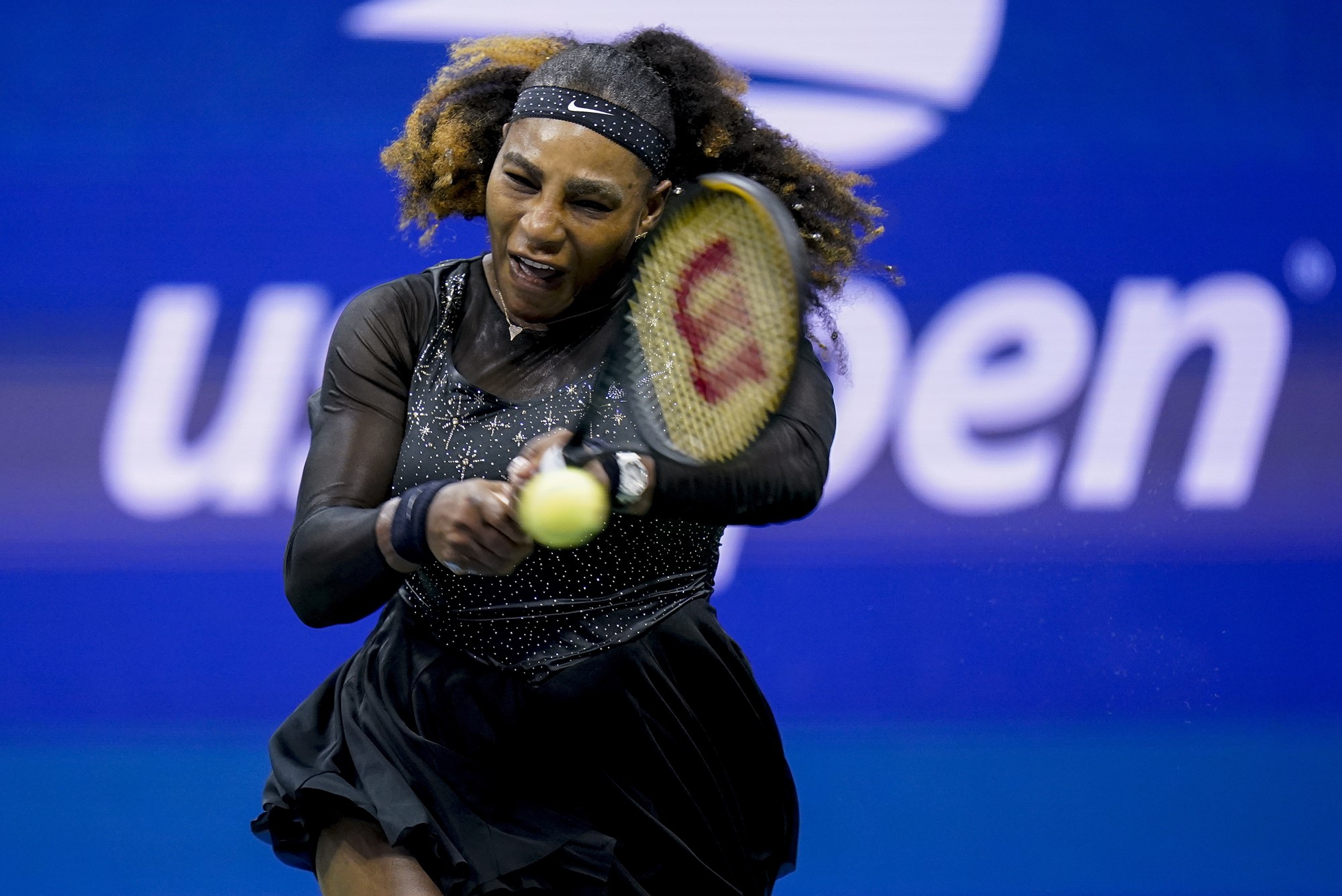 A woman in all black hits a tennis ball with a raquet.