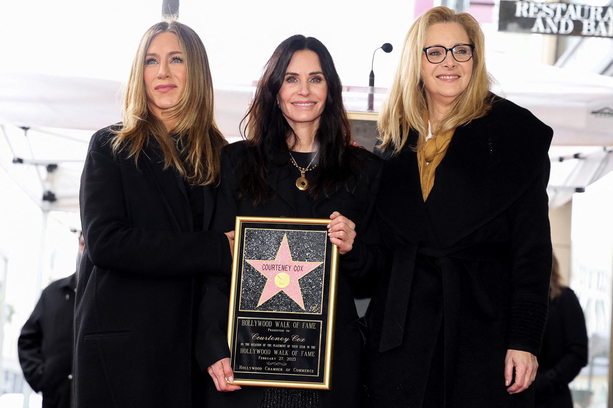Jennifer Anniston, Courtney Cox and Lisa Kudrow stand holding a Hollywood Walk of Fame plaque.