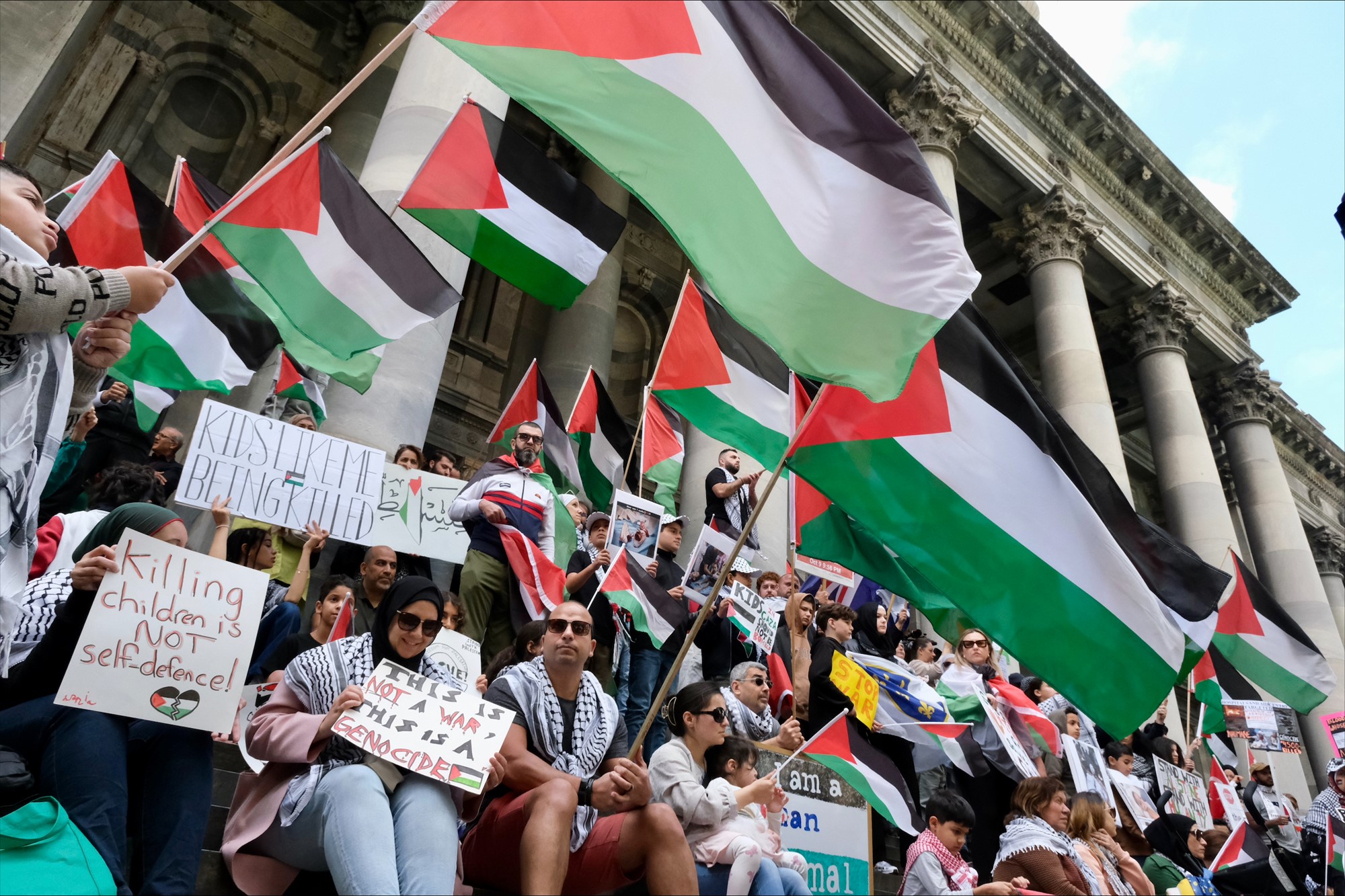 A large group of people carrying signs and Palestinian flags sit on the steps of a stone building.