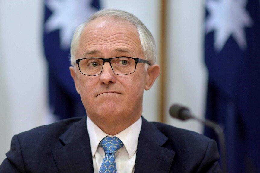 Malcolm Turnbull sits at a microphone, wearing a suit and glasses, with Australian flags in the background