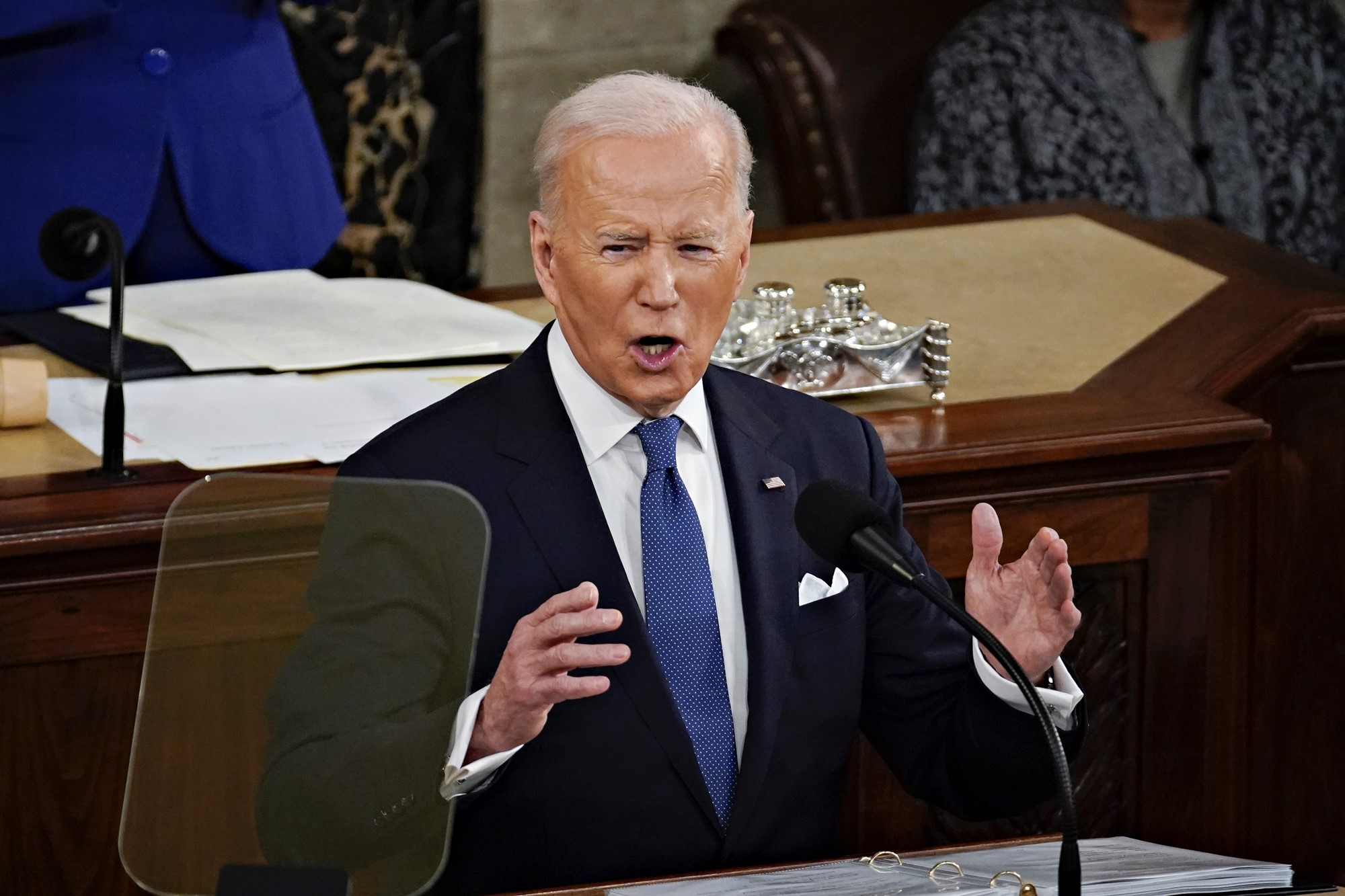 US President Joe Biden  stands at a desk with a microphone. His hands are raised. 