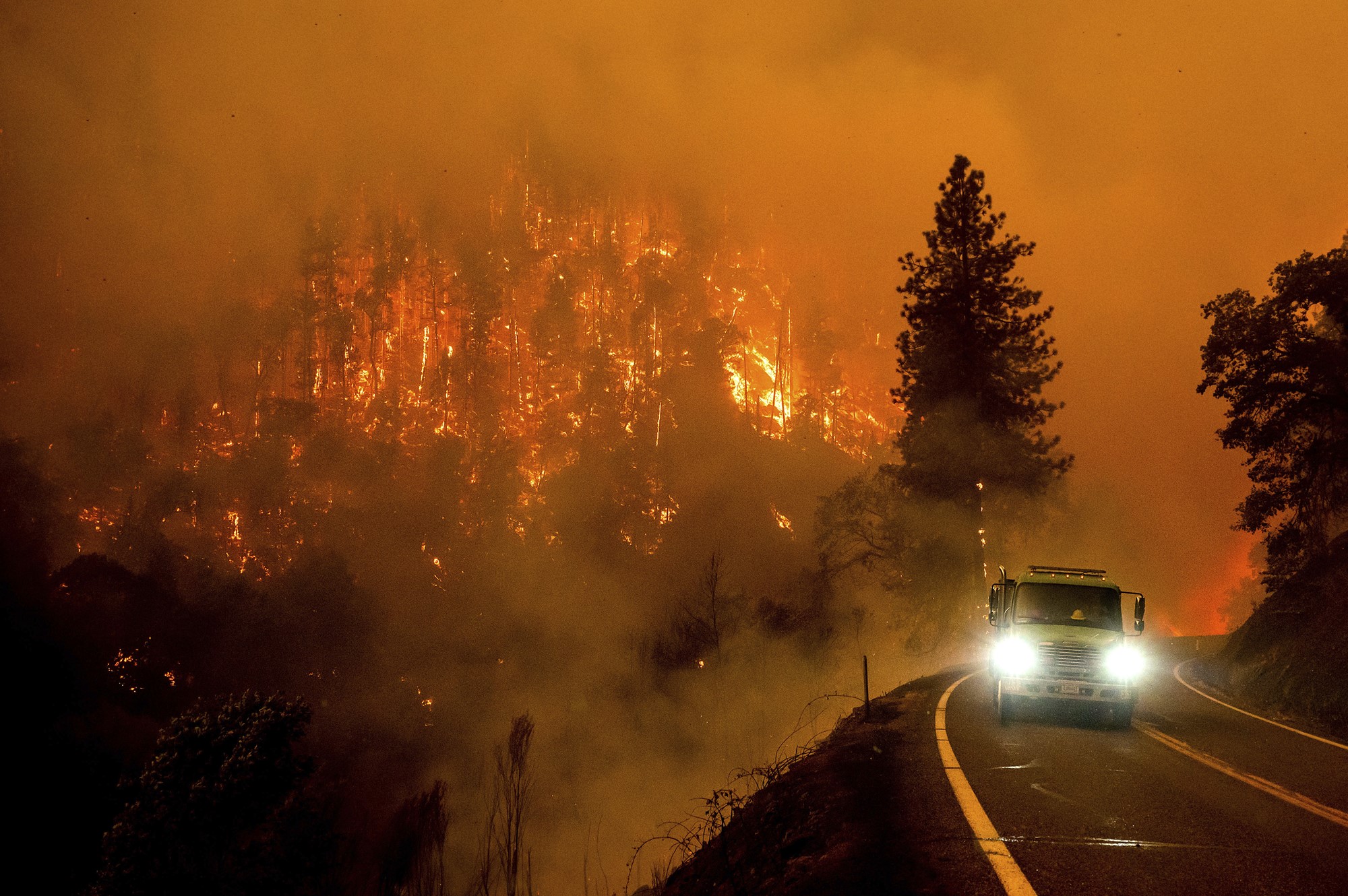 A firetruck drives along a road with burning trees and a glowing orange sky in the background.