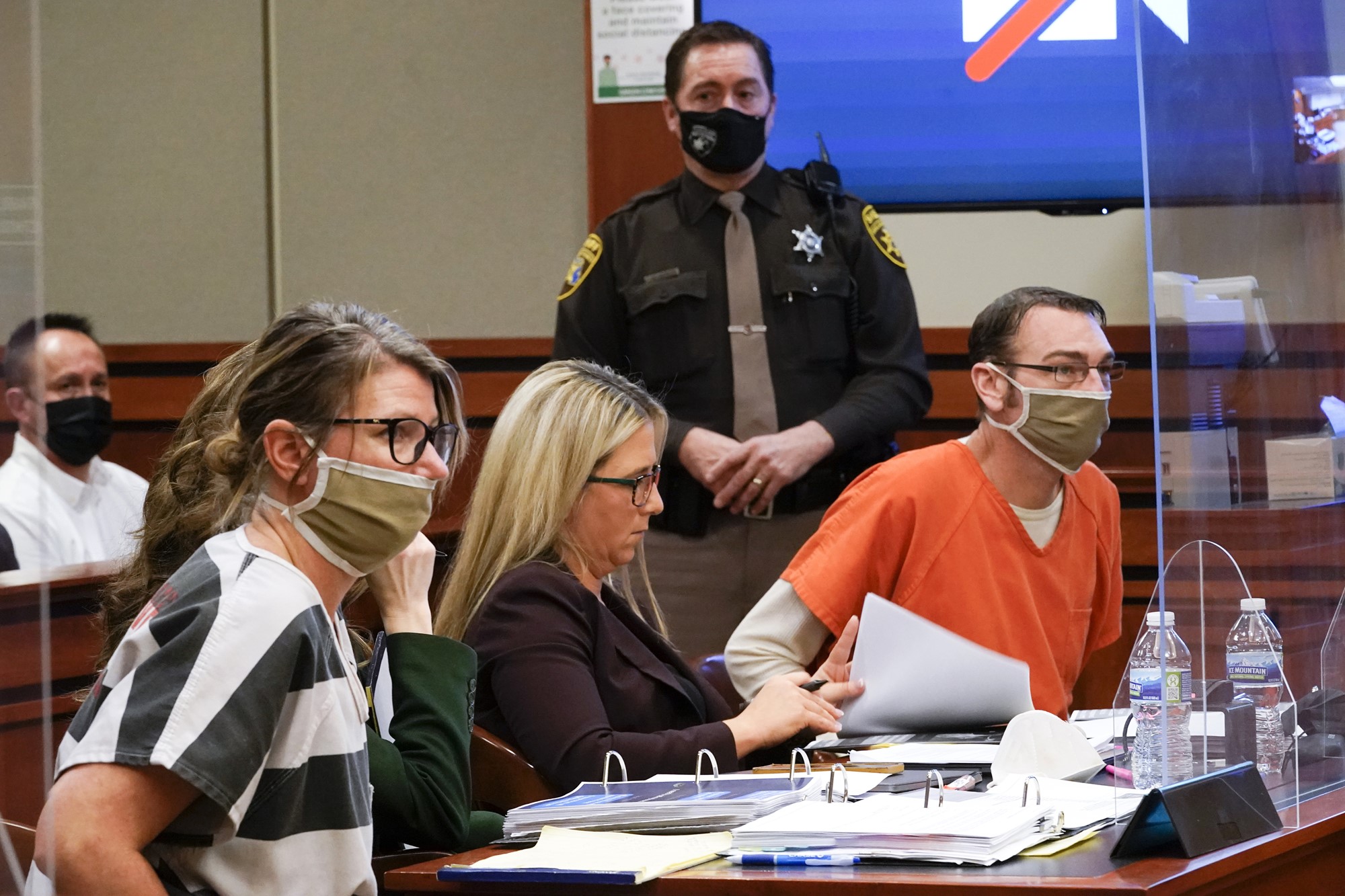 A woman and a man wear prison outfits, sitting at a desk in a courtroom.