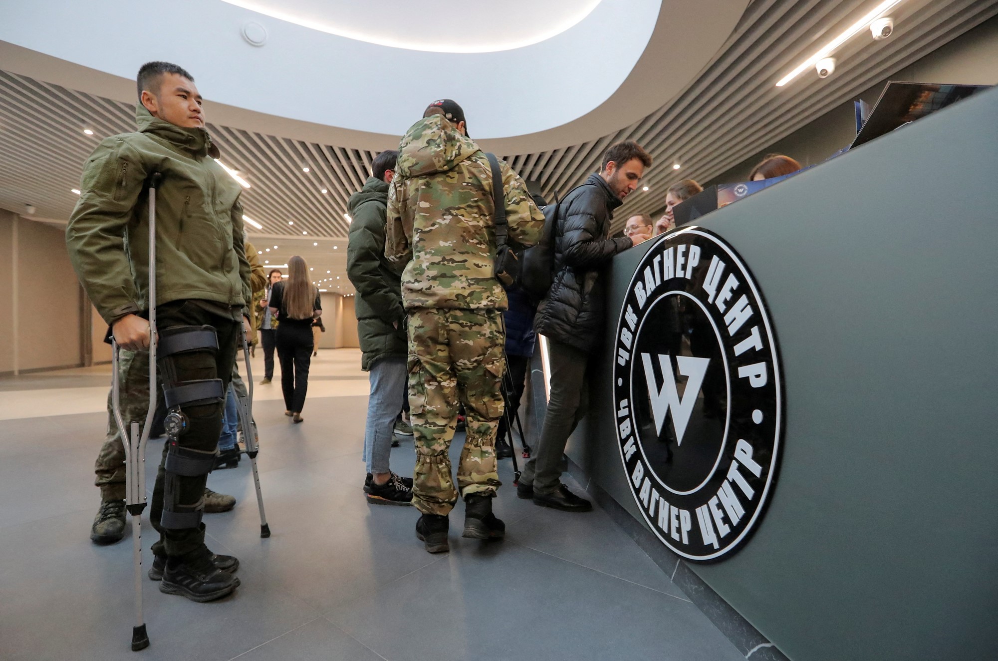 Men in camouflage and one on crutches stand at the front desk of a building with the Wagner logo