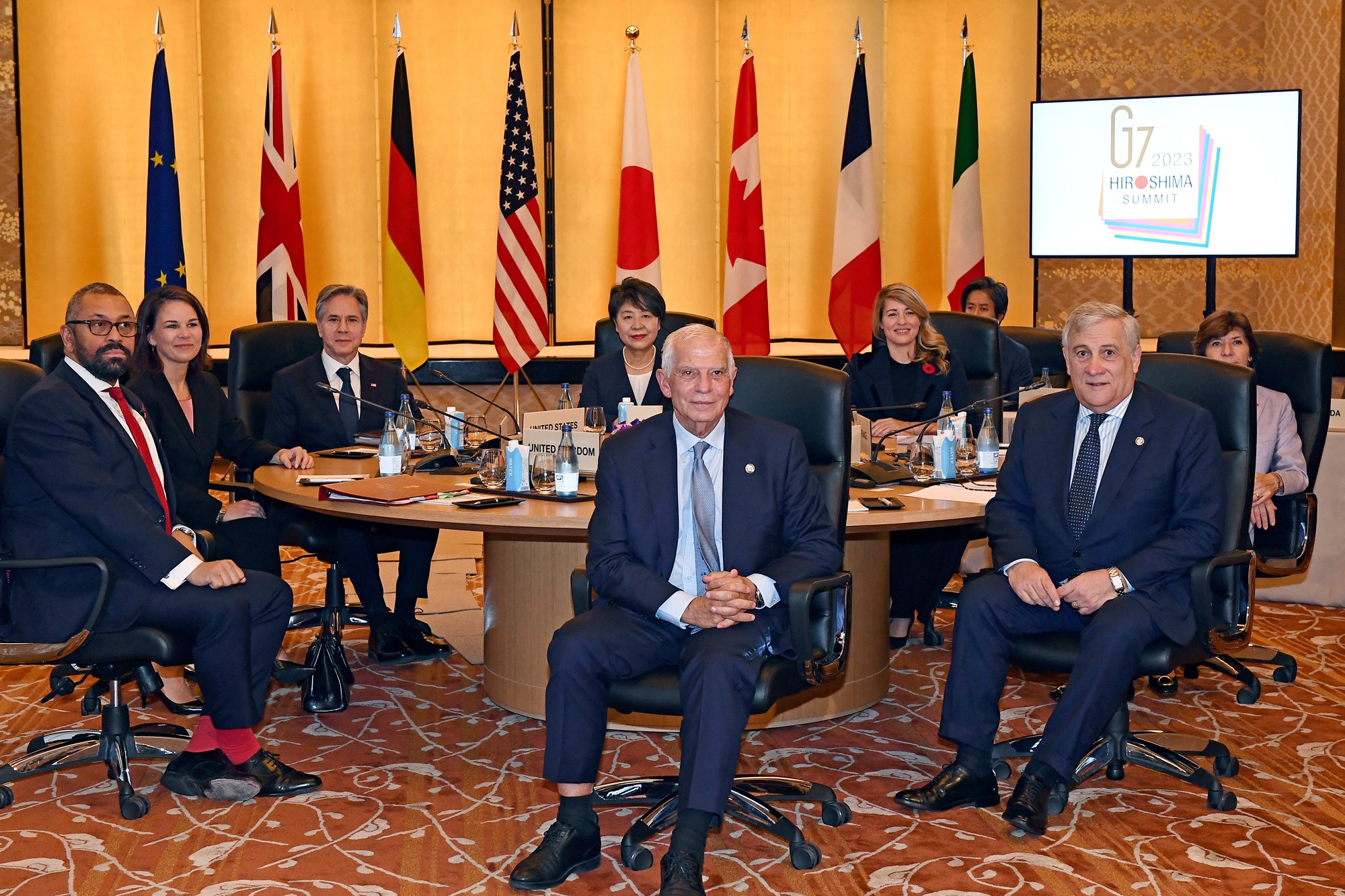 G7 prime ministers sit around a round meetng table, with the countries flags on stands in the background