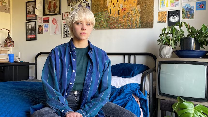 A young person with short blonde hair sits on a bed next to a CRT TV with plants on it, and artwork on the wall behind them
