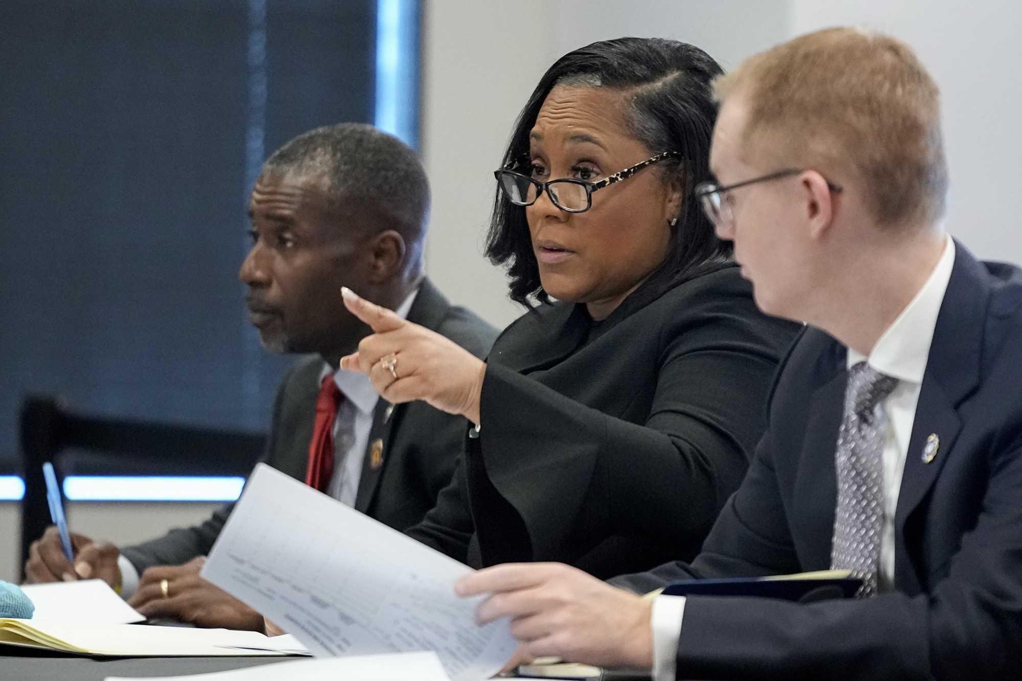 A black woman in glasses and a suit sits between two men at a table, speaking and pointing into a room